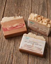 Three artisanal handcrafted soap bars from RedC Essentials, including Gingerbread Cookies and Marbled Marble, arranged on a rustic wooden surface.