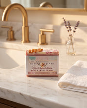 A rectangular bar of soap labeled 'Rose Clay & Honey' by Red Oak Essentials sits on a white marble bathroom counter with gold faucets, a folded white towel, and dried lavender in the background.
