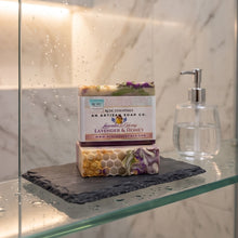A bar of lavender and honey scented soap by An Artisan Soap Co. is displayed on a glass shelf inside a shower enclosure, next to a clear glass pump bottle.