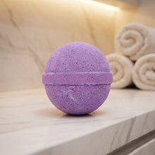 A purple spherical bath bomb with a horizontal seam, resting on a light marble countertop with rolled towels and a warm light fixture in the background.