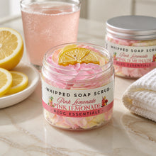 Jars of whipped soap sugar scrub with pink and yellow colors, alongside slices of lemon and pink flowers, indicating a citrus scent profile.