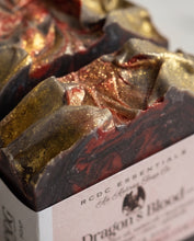 A close-up of a handcrafted soap bar with a marbled design of dark brown, metallic gold, and deep red, resting in a white paper box labeled 'RCDC ESSENTIALS' and 'Dragon's Blood'.
