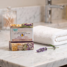 Two stacked bars of artisan soap from Lavender & Honey by An Artisan Soap Co. sit on a white marble bathroom counter next to a folded white towel and a sprig of lavender, with a blurred sink and faucet in the background.