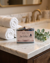 A bar of goat's milk soap from 'The Perfect Man' by 'MCDC Essentials' sits on a white marble bathroom counter next to two rolled white towels and a green eucalyptus sprig, with a blurred gold faucet and mirror in the background.