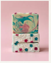 Three stacked rectangular bars of handmade soap on a solid pink background. The top bar features a marbled swirl pattern of teal, pink, and cream.