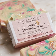 A bar of handmade soap labeled 'RCDC Essentials Sweet Honeysuckle' sits on a marble-like surface with other marbled soap bars around it. The main soap bar has a white paper wrapper with pink and black text, and the soap itself shows pink, mint green, cream, and gold marbling.