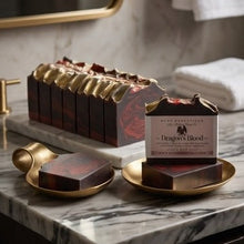 A set of dark red and gold artisanal soap bars displayed on a marble bathroom counter, including a boxed bar labeled 'Dragon's Blood' by Blue Mountain Soap.