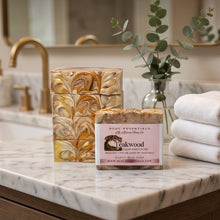 Two bars of handcrafted soap with a marbled pattern in beige, brown, and gold, displayed on a white marble bathroom counter next to a glass vase with eucalyptus and folded white towels. The front bar has a pink label reading 'MCC Essentials The Alchemy Soap Co.