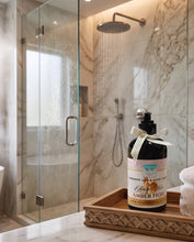 A bottle of Shower Gel & Body Wash labeled 'Amber Noir' by 'Scent of Luxury' sits on a carved wooden tray in a modern marble bathroom next to a glass shower enclosure with water running from a round shower head.
