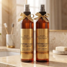 Two amber spray bottles from RCDC Essentials 'Regina Collection' labeled 'Fall Into Cozy' body linen spray and room linen spray, tied with gold ribbons, displayed on a marble bathroom counter.