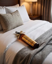 A luxury bottle of hair product tied with a beige ribbon rests on a neatly made bed with white linens and a gray throw blanket in a warm, softly lit bedroom.