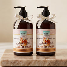Two amber pump bottles of RCDC Essentials Shower Gel & Body Wash in Amber Noir, tied with cream ribbons, displayed on a marble slab against a warm beige background.