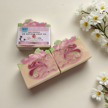 Two bars of soap with pink and green swirls next to their packaging on a light surface with flowers.