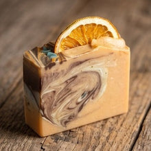 A rectangular bar of artisanal soap with a marbled swirl pattern in shades of orange, brown, and cream, topped with a dried orange slice, set on a rustic wooden surface.