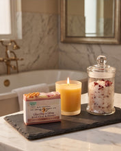 A styled bathroom scene with a bar of soap, a lit candle, and a glass jar of bath salts arranged on a dark tray on a marble countertop, with a bathtub and mirror in the background.