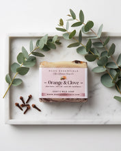 A rectangular bar of soap labeled 'Orange & Clove' by HCCD Essentials is arranged on a white marble tray with eucalyptus leaves and cloves. The soap has a pale pink label and a rustic tan top layer.