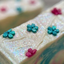 A close-up of a rectangular bar of soap with an iridescent white surface decorated with small blue and pink flower motifs, with a second soap bar blurred in the background.