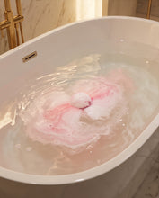 A white freestanding bathtub filled with water, showing a pink and white bath bomb or bath foam burst in the center. Gold-tone faucet hardware is visible against a marble bathroom wall and floor.