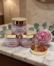 Three jars of Whispered Soap Scrubs Woodstock Whipped Soap Scrub with gold lids are arranged on a marble countertop, alongside a pink rose-shaped bath bomb and its gold lid, with green leaves in the background.