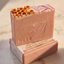 A handmade soap bar in a dusty pink color with a swirled purple pattern and a honeycomb design on top, displayed on a marble surface with its branded paper packaging box.