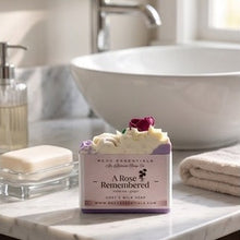 A bar of goat's milk soap labeled 'A Rose Remembered' by Milk Essentials, styled on a marble bathroom counter with a white vessel sink, folded towel, and soap dispenser in the background.