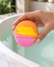 A hand holding a two-toned spherical bath bomb with a pink bottom half and an orange top half separated by a yellow band, positioned over a bathtub filled with water.