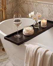 A dark wooden bath tray draped over the side of a white freestanding bathtub, styled with a wine glass, a candle, a small flower vase, and a jar of body butter in a marble bathroom setting.