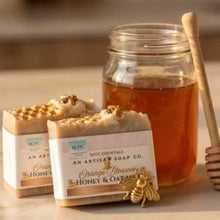 Two bars of handmade honey and oatmeal soap with gold foil flakes and a wooden honey dipper in a glass jar, set on a light marble surface with a warm, blurred background.