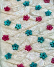 A close-up of a textured cream-colored fabric with a crisscross pattern, decorated with small glittery flower motifs in teal and magenta pink.