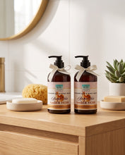Two amber pump bottles of WCOF Flower Power & Body Wash in Amber Noir scent, styled on a wooden bathroom counter with soap dishes, a sponge, and a small succulent.
