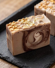 A rectangular bar of handmade soap with a brown and beige swirl pattern and gold leaf flakes on top, displayed on a black slate tray.