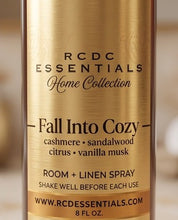 A close-up of a cylindrical bottle labeled RCDC Essentials Home Collection 'Fall Into Cozy' room and linen spray, with a brushed gold label and black text on a softly blurred warm background.