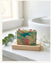A rectangular bar of marbled soap with swirls of green, yellow, pink, and blue sits on a small wooden soap dish on a white marble bathroom counter, with a white sink and tiled wall in the background.