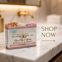 A bar of soap from Redc Essentials An Artisan Soap Co. labeled 'Rose Clay & Honey' sits on a marble countertop in a warm, softly blurred bathroom setting.