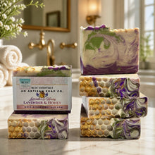 Four artisanal lavender and honey soap bars with gold foil honeycomb patterns and purple swirls, with one wrapped in a paper label. The soaps are arranged on a marble countertop in a warm, softly-lit bathroom setting with gold fixtures and rolled towels in the background.