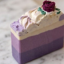 A rectangular layered soap bar with purple and cream bands, topped with swirled white frosting and a small dark purple rose decoration, shown on a light marble-like surface.