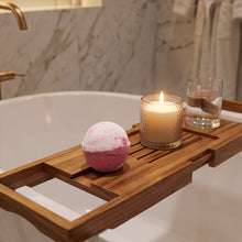 A wooden bath caddy draped over a white bathtub, holding a pink and white bath bomb, a lit candle in a textured glass holder, and a glass of water. The background shows a marble-tiled bathroom wall and a robe.
