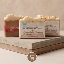 Three artisanal gingerbread cookie soap bars with cream-colored whipped tops and pink/red marbled bodies, displayed on a wooden block with a concrete top. The center bar is in front with a label reading 'RCDC Essentials An Artisan Soap Co.