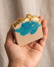 A hand holding a rectangular bar of handmade soap with a beige base, bright blue swirled pattern, and a textured gold leaf design on top, set against a soft beige fabric background.