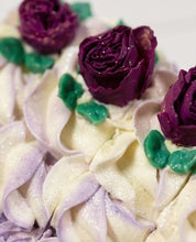 A close-up of a decorated cake or pastry with piped cream-colored frosting, purple rose decorations, and green leaf accents.