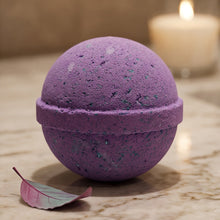 A purple spherical bath bomb with subtle blue-green glitter flecks, resting on a marble surface next to a small pink leaf, with a softly blurred lit candle in the background.