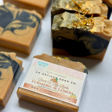 A close-up of artisanal soap bars with a paper label reading 'Greek To My Roots Turmeric & Lemon' by Rutie Essentials, shown on a white surface with additional marbled soap bars in the background.