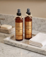 Two amber spray bottles labeled 'Bodc Essentials Stay Awkward' with beige ribbons, standing on a marble tray beside a folded white towel and soap dish in a bathroom setting.