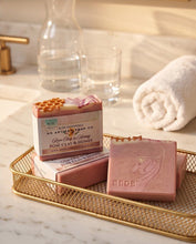 Three pink artisan soap bars displayed on a gold wire tray on a marble surface, with a glass bottle, glass, and rolled towel in the background.