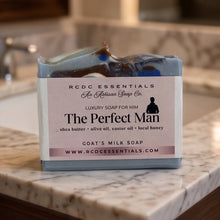 A bar of RCDC Essentials 'The Perfect Man' luxury soap, featuring a light blue and white marbled body with dark brown and blue inclusions, wrapped in a white paper label and displayed on a marble countertop.