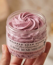 A hand holding a clear jar of whipped soap scrub with a pink-lilac swirled scrub inside. The label reads 'Whipped Soap Scrub' and 'A Rose Remembered' with the brand 'PCC Essentials' visible.