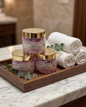 Three jars of whipped soap scrub by Woodstock Apothecary arranged on a wooden tray with rolled white towels and greenery, set on a marble countertop in a spa-like setting.