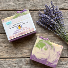 Lavender and Honey artisan soap bar with purple and green swirls, shown next to its paper packaging box and a sprig of dried lavender on a rustic wooden surface.