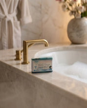 A bar of artisan soap labeled 'Sapio Sumo' sits on the edge of a marble bathroom sink next to a brushed gold faucet, with a blurred warm-toned bathroom background.