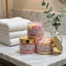 Three jars of whipped soap scrub by Sweet Honeysuckle Bath & Body Products arranged on a white marble bathroom counter next to folded white towels and a glass vase with greenery.